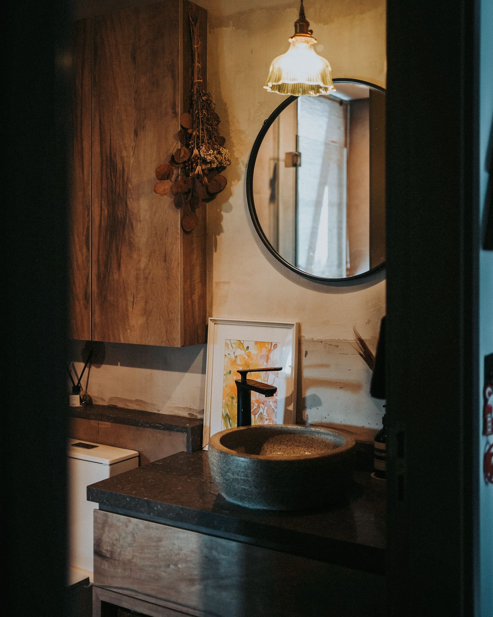 Nostalgia meets postmodernism - Wooden cabinetry, rustic design, hanging dried plants, round mirror, framed artwork, stone
