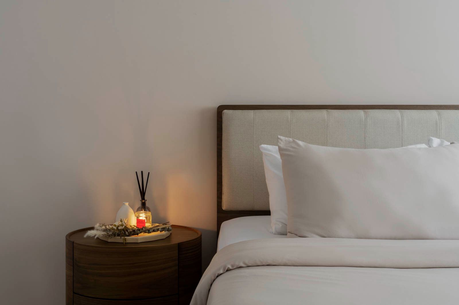 Our master bedroom is kept to the necessities - Wooden bedside table, white pillows, beige headboard, soft lighting, serene
