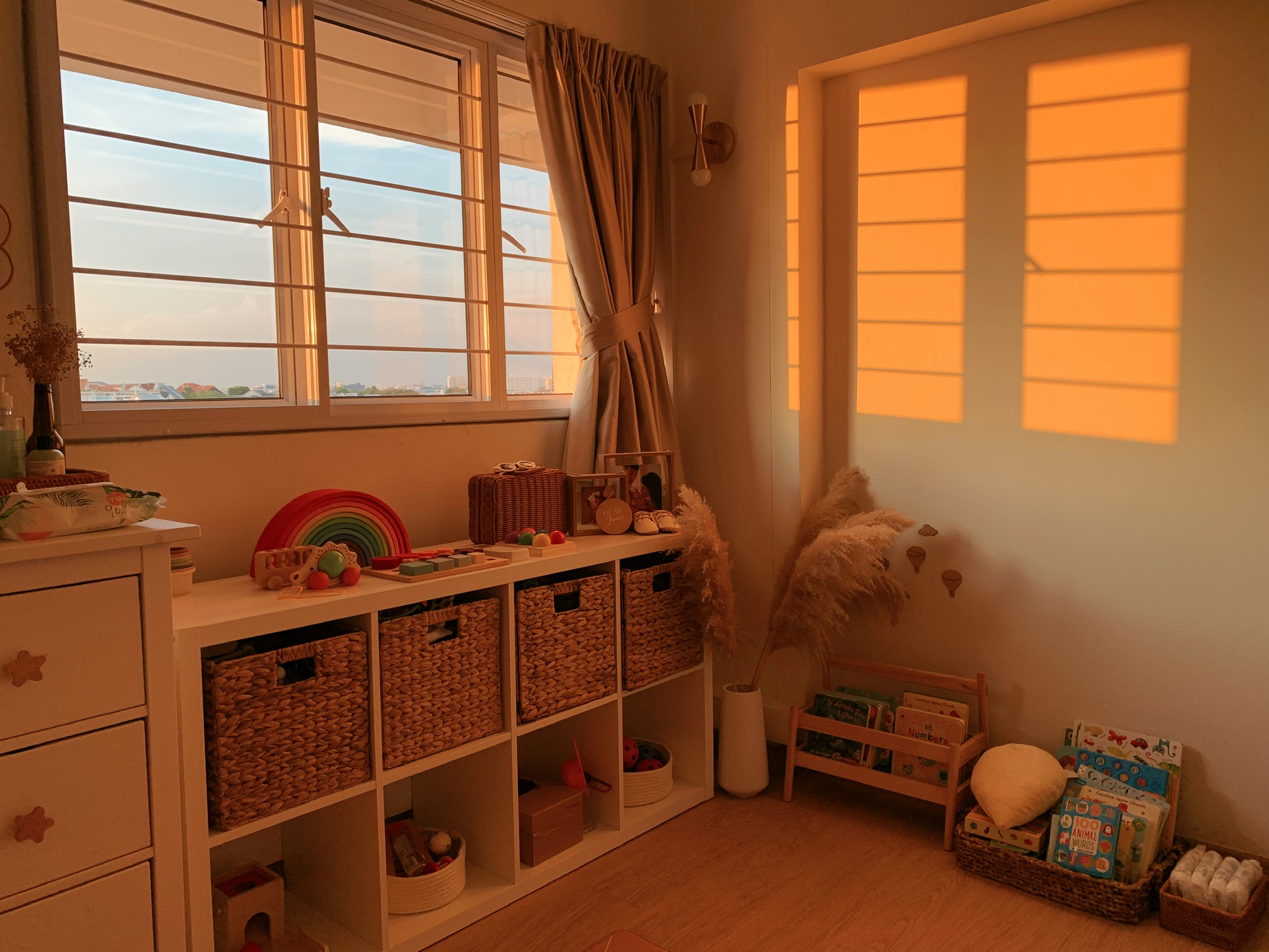 Our warm nursery - Sunset view through large windows, warm lighting, soft pastel colors, cozy room ambiance, children's toys