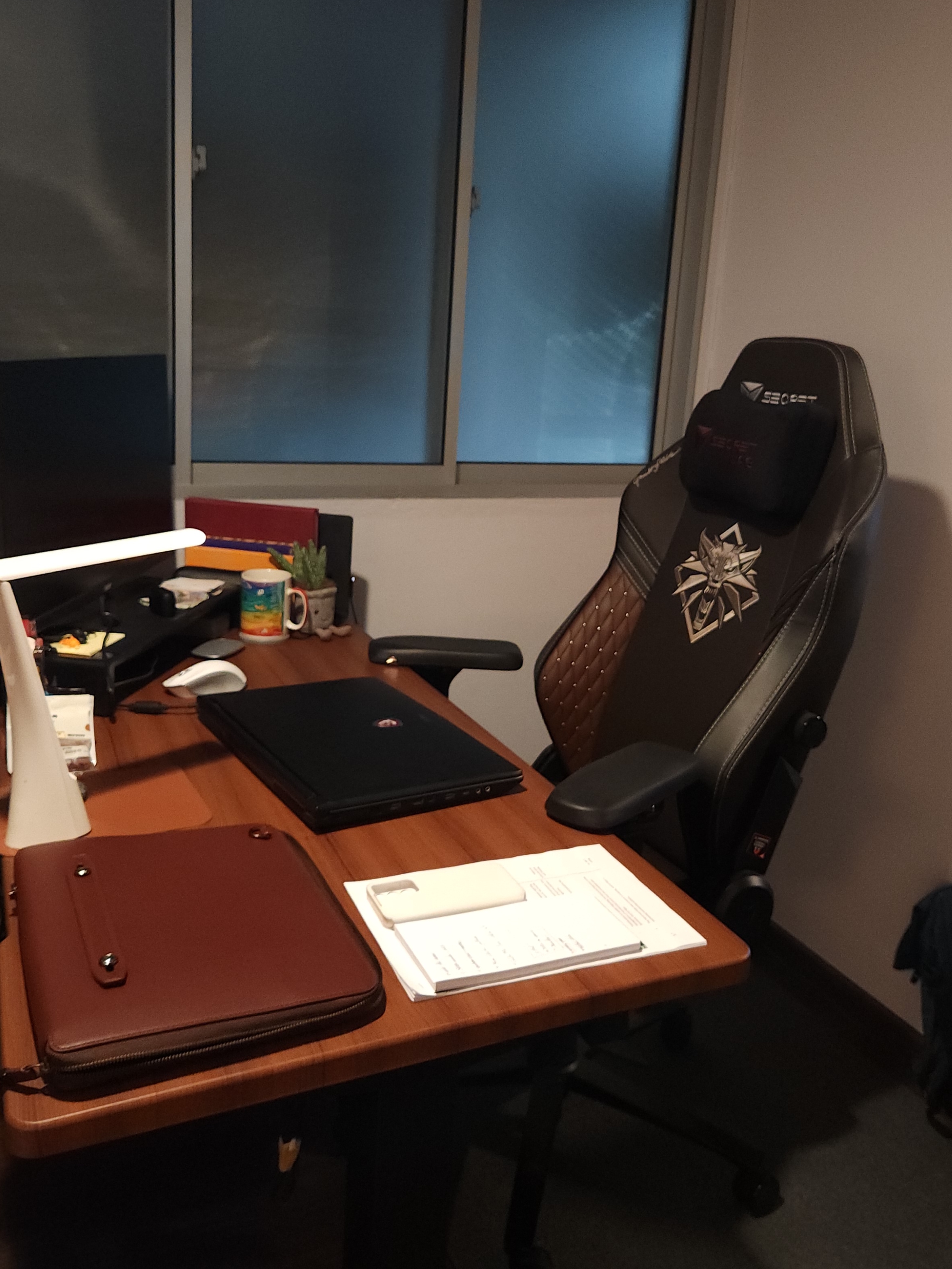 Our Trapezoidal Glass Office Where We WFH Everyday - Black gaming chair with "Secret" logo, brown leather laptop, white desk