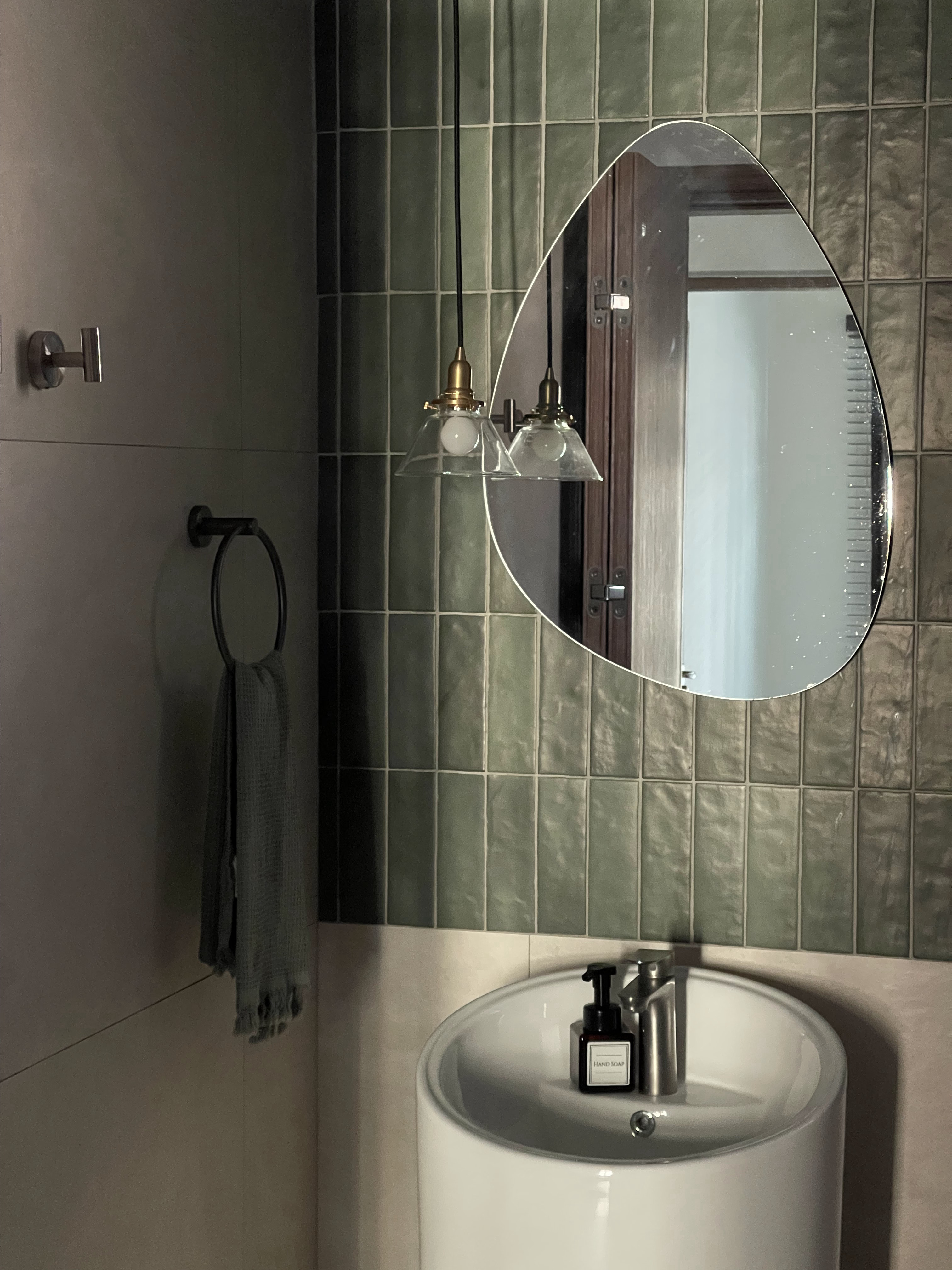 Trunks and Trees Inspired Home - Green tiled wall, round mirror, hanging light fixture, white basin, hand soap bottle
