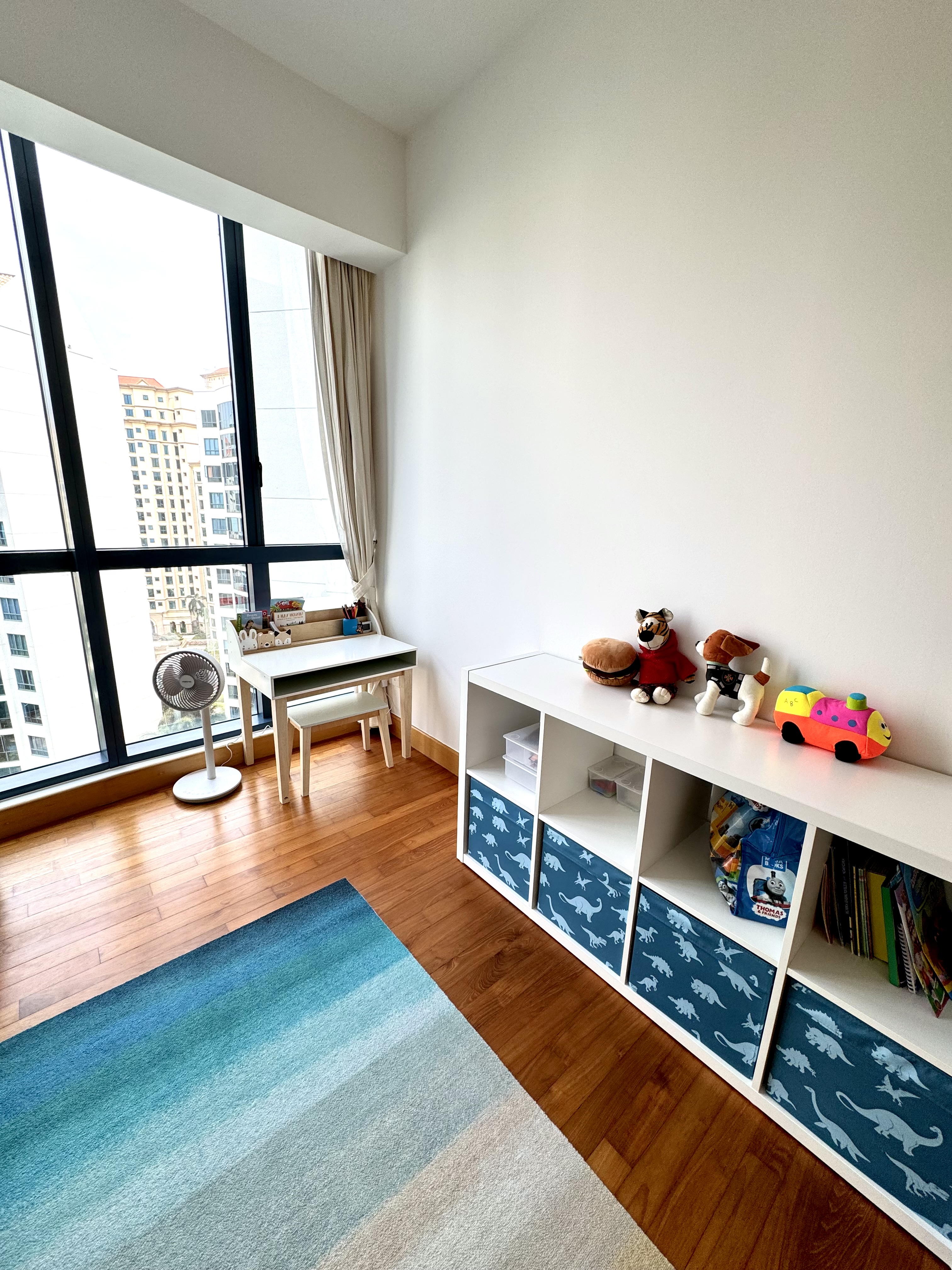 Our child’s room  - Large windows, beige curtains, wooden floor, blue rug, white shelf, dinosaur-themed storage, colorful