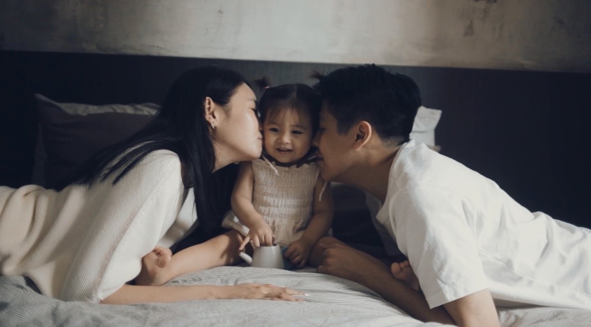 A place to rest and recharge  - Mom and Dad kissing baby on bed, loving family moment, tender interaction, warm embrace