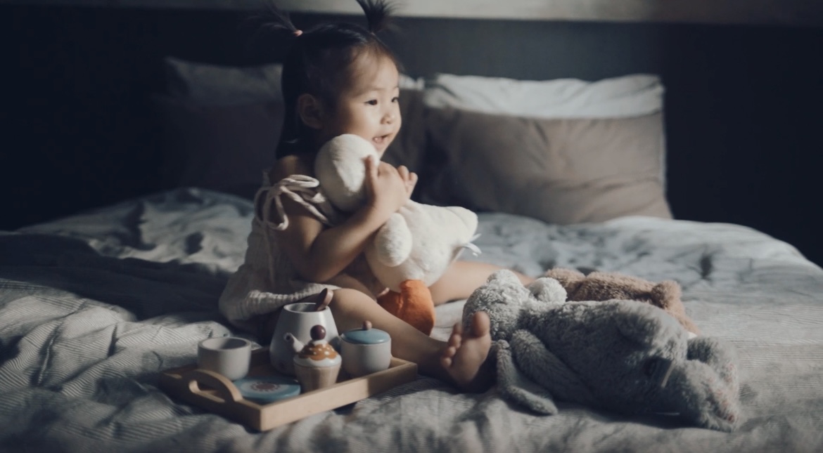A place to rest and recharge  - Soft, cozy bedroom setting, child sitting on bed, holding a plush toy, gray and white