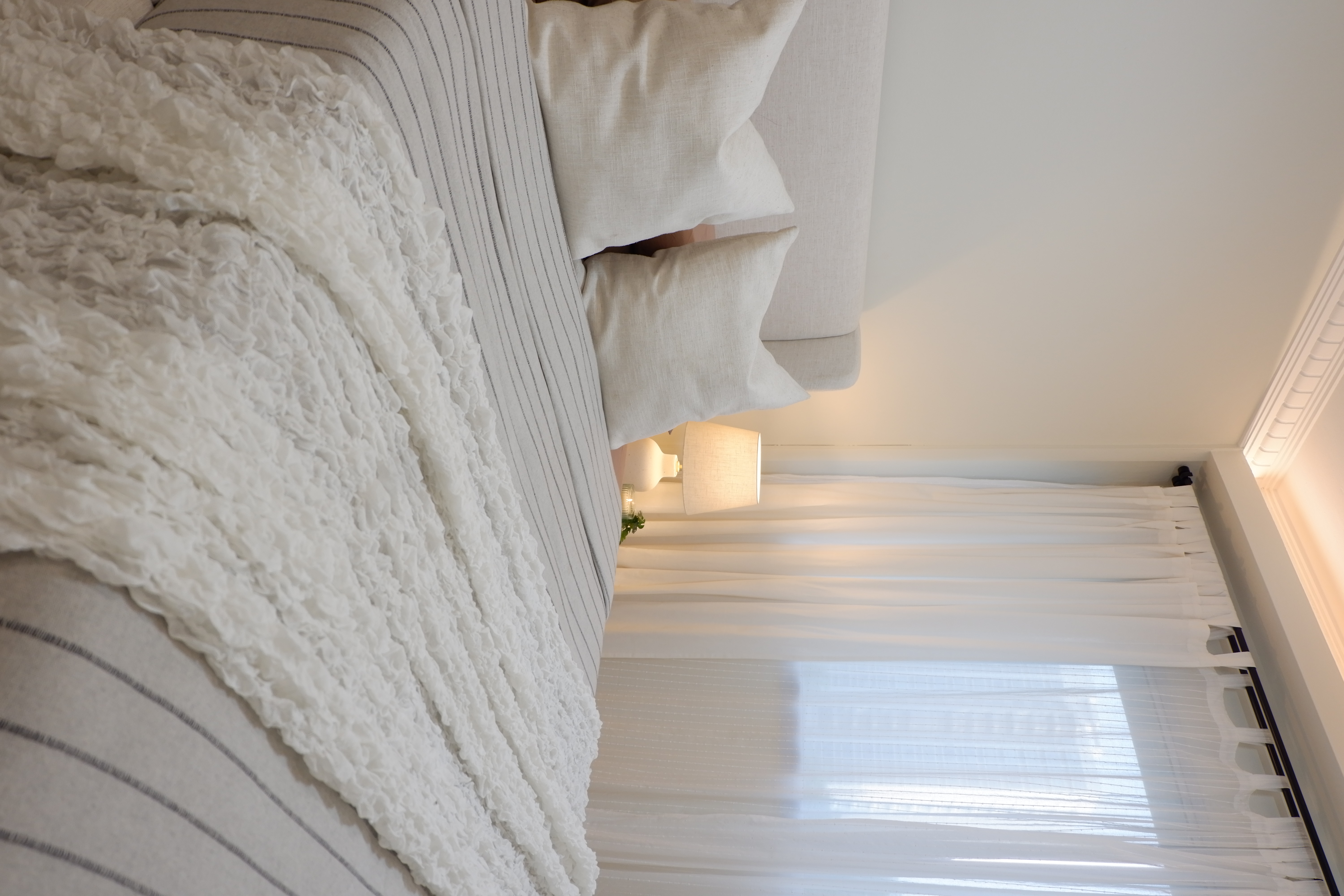Warm and Bright Bedroom Retreat - White textured bedding, beige pillows, soft lighting, window with sheer curtains, cozy room