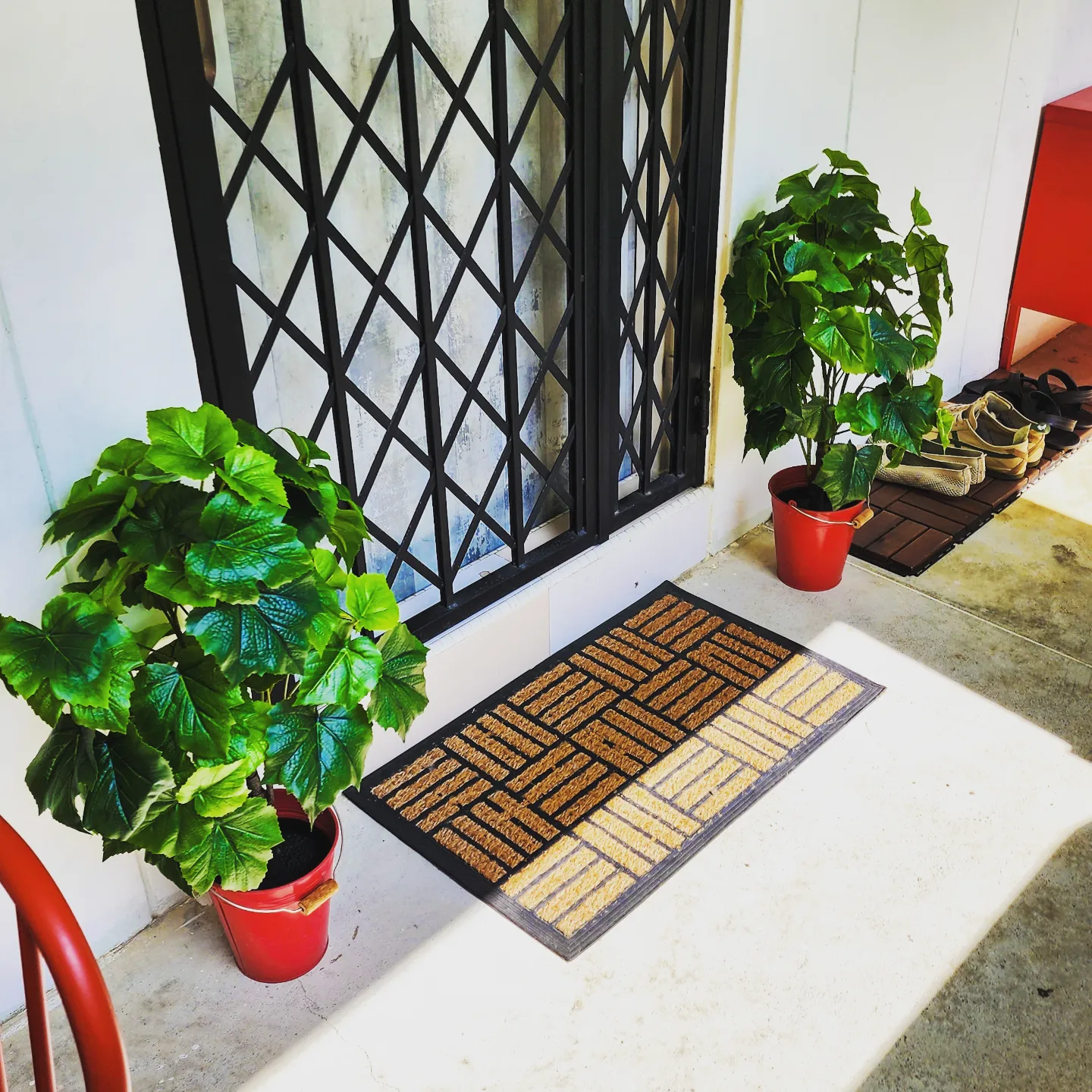 Our Zen corner - Green potted plant, red watering can, black metal gate, white wall, brown doormat, green leaves