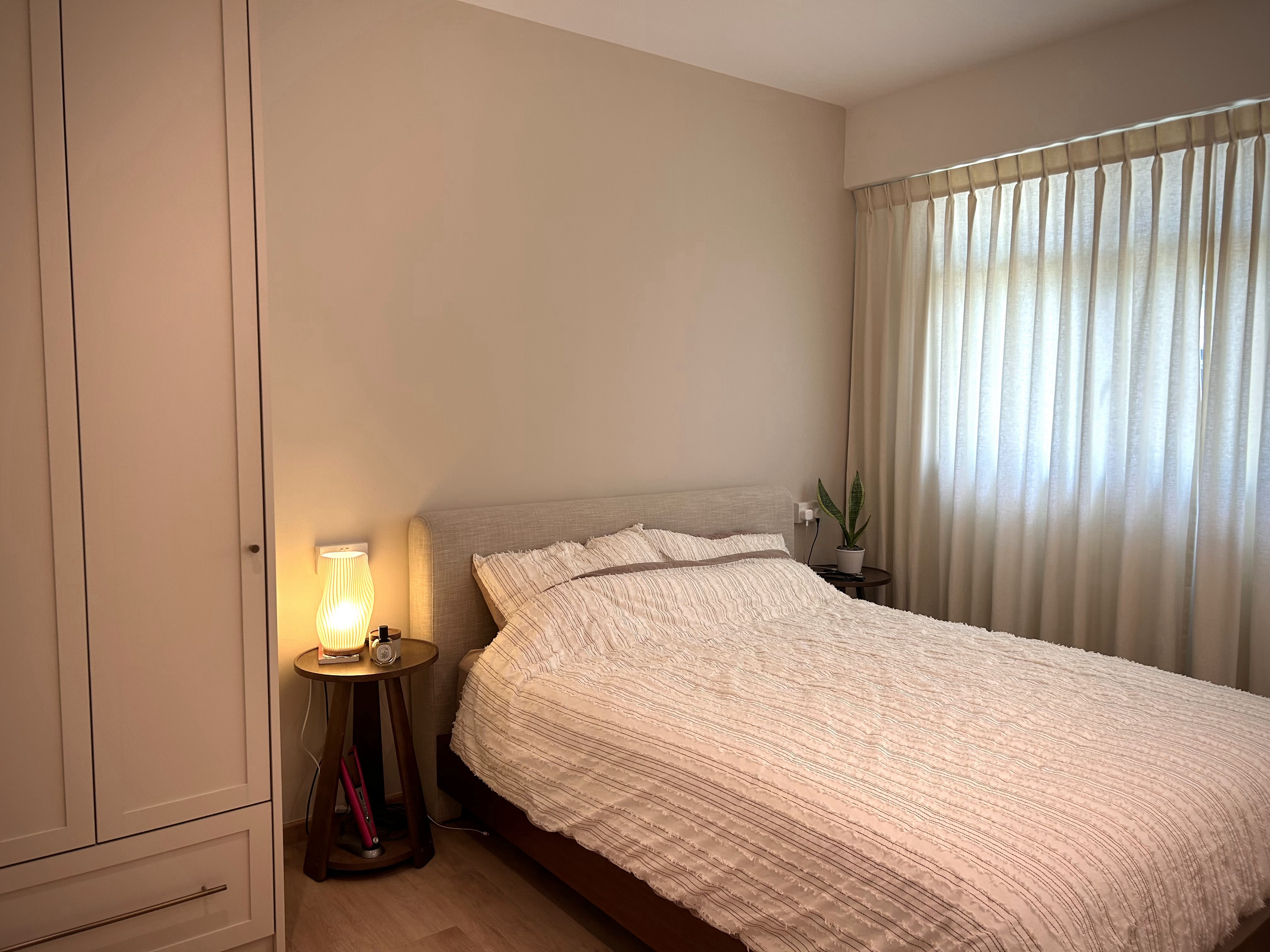 A Peek into Our Cozy Bedroom! - Warmly lit bedside table, soft beige curtains, cozy bed with white and brown striped