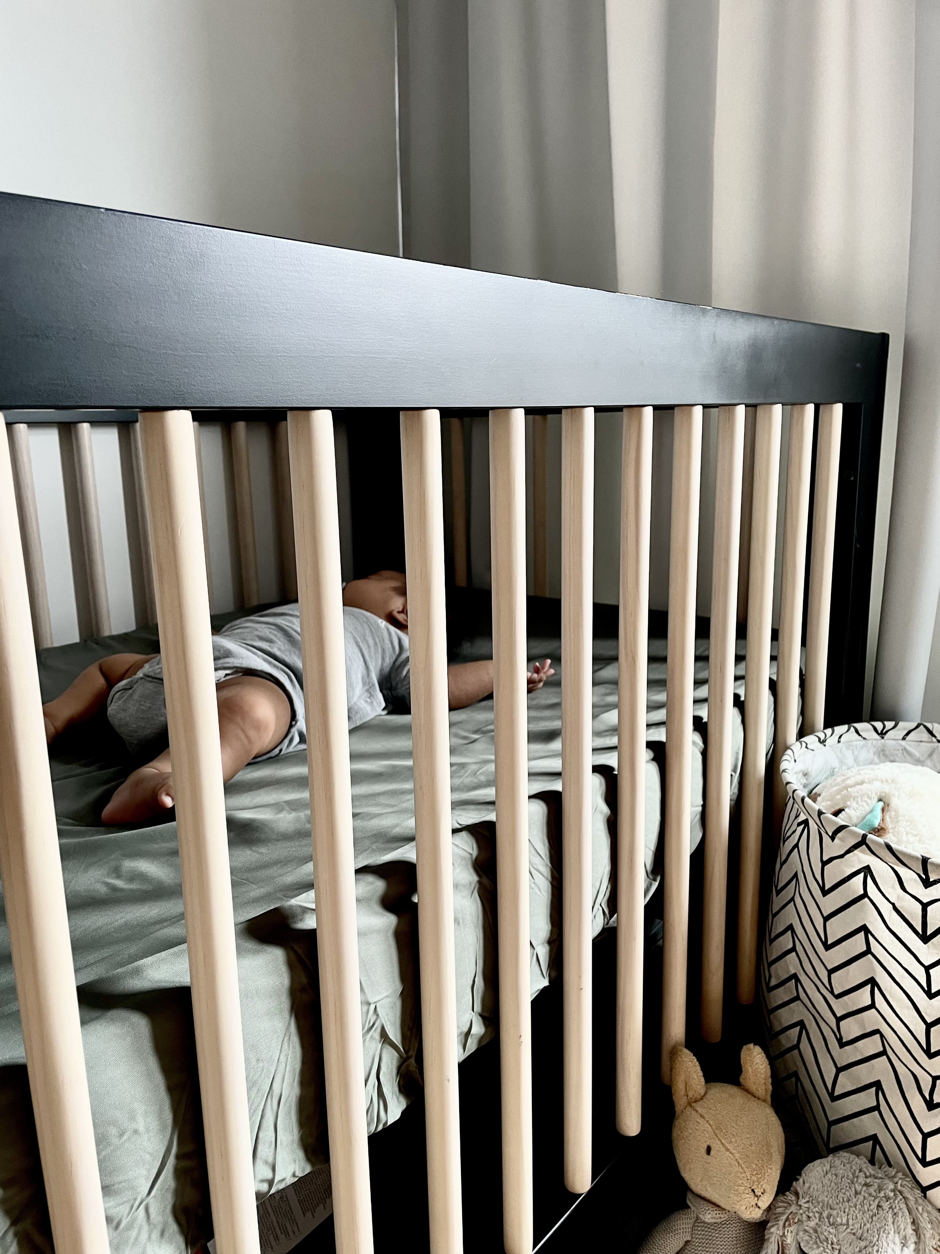 How I decorate my baby’s room - Wooden crib, baby lying in bed, gray bedding, wooden bars, baby's legs visible, beige