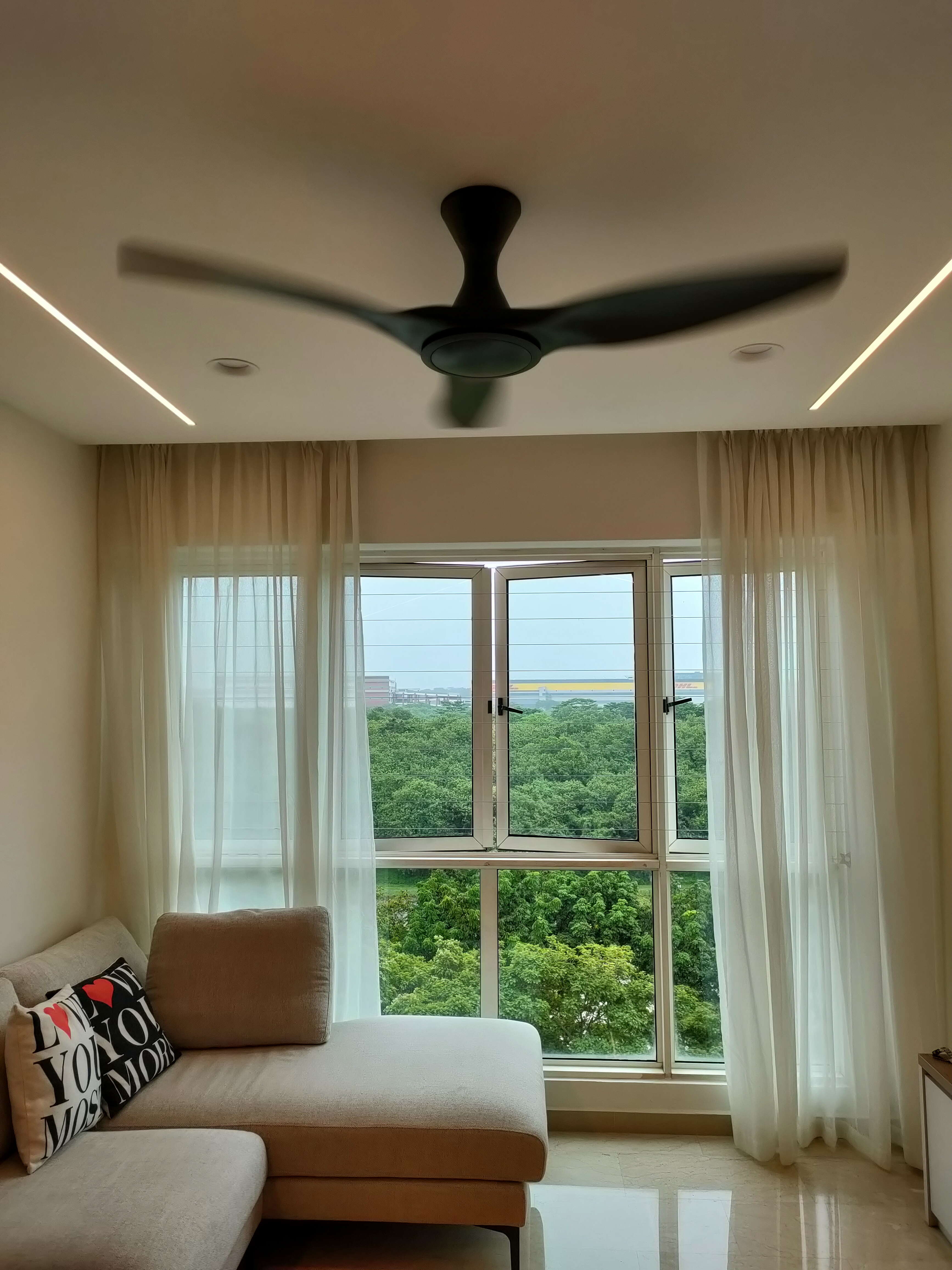 What makes the house our home... - Modern living room, ceiling fan, beige walls, large window, sheer curtains, natural light