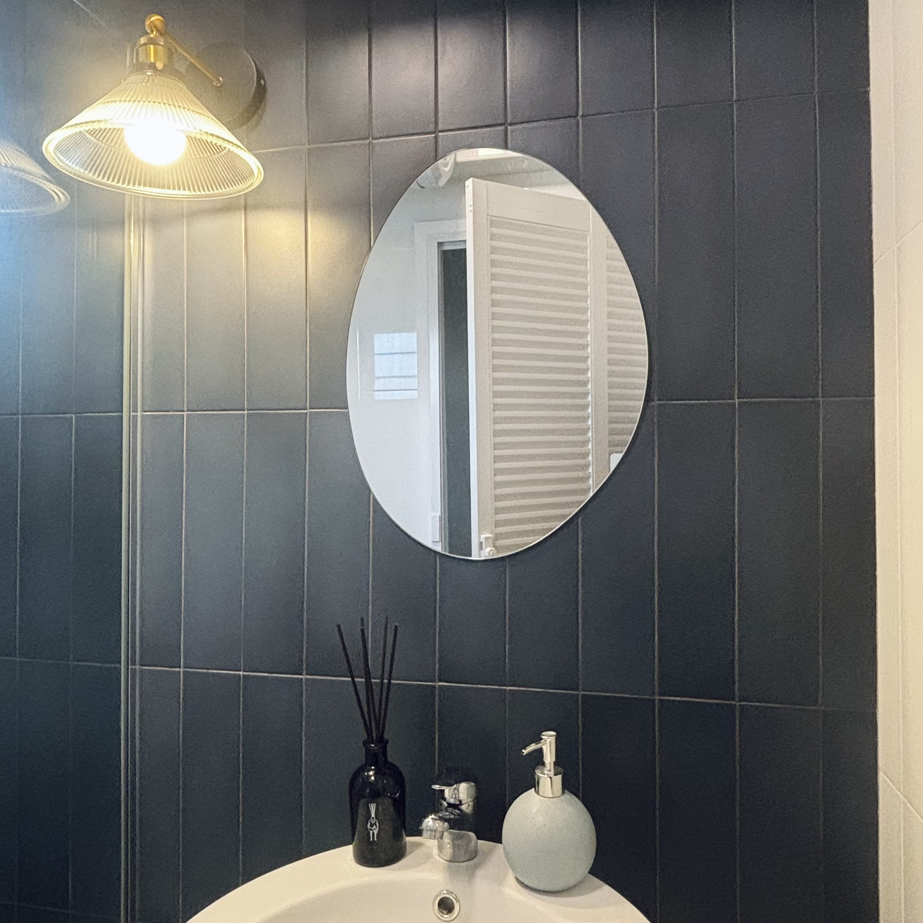 Two toilets, two different themes ✨ - Round mirror, dark tiled wall, white sink, black vase, light fixture, open door