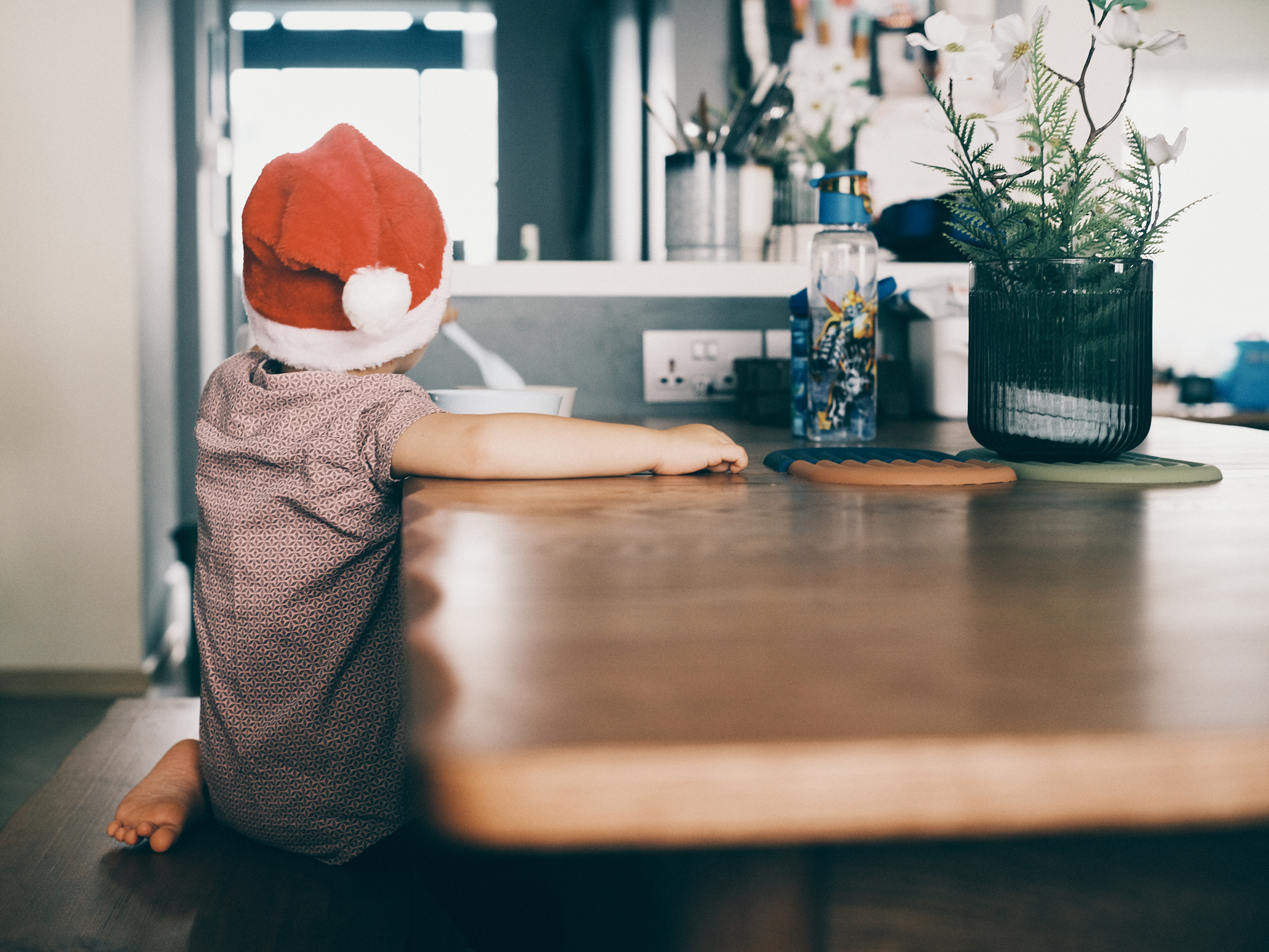A mid-century family dining area - Santa hat, child, sitting, wooden table, patterned shirt, white socks, green plant, blue