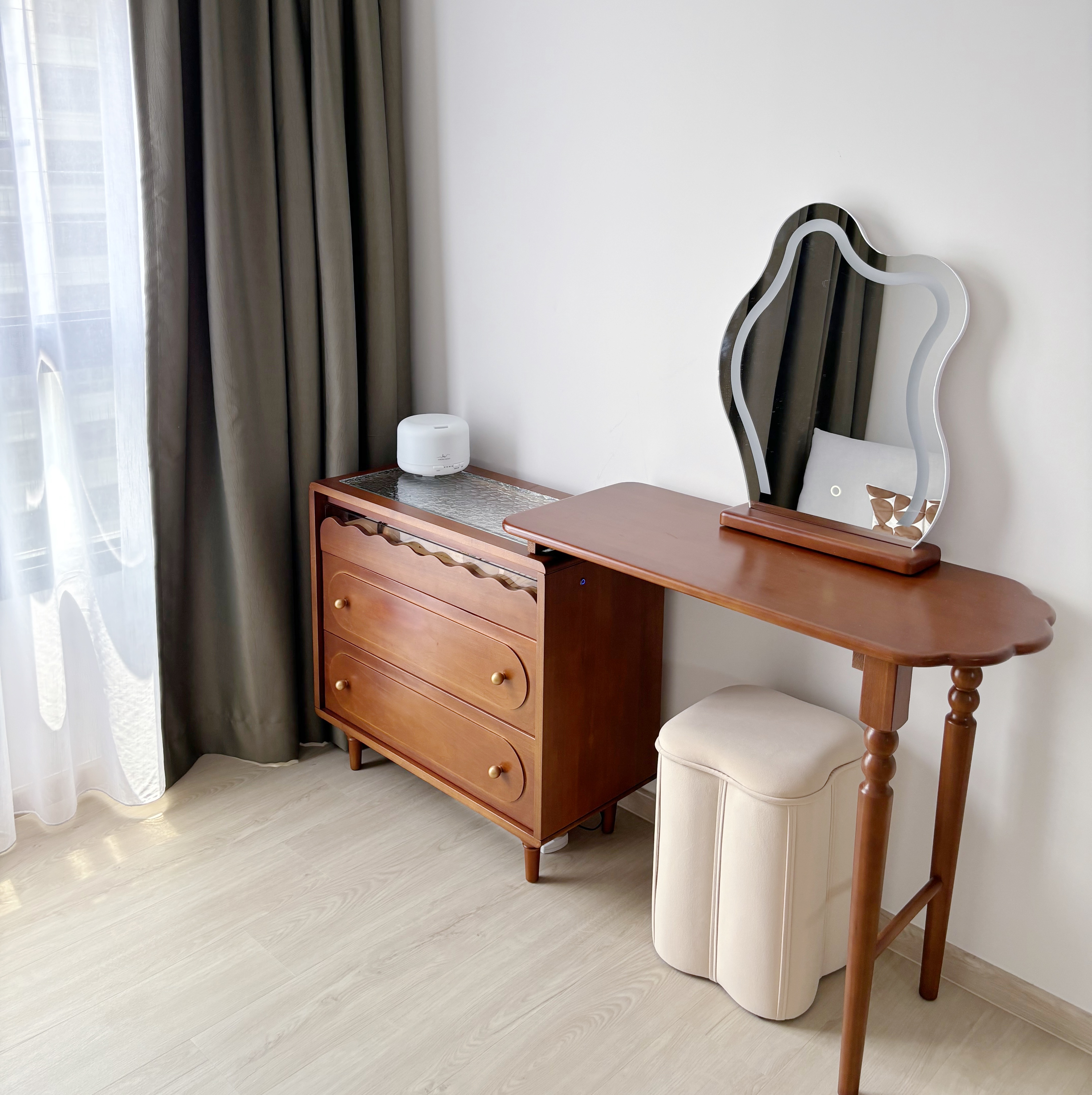 Humble Cozy Guest Bedroom - Wooden dresser with drawers, white lamp on top, mirror on table, beige ottoman, light wooden
