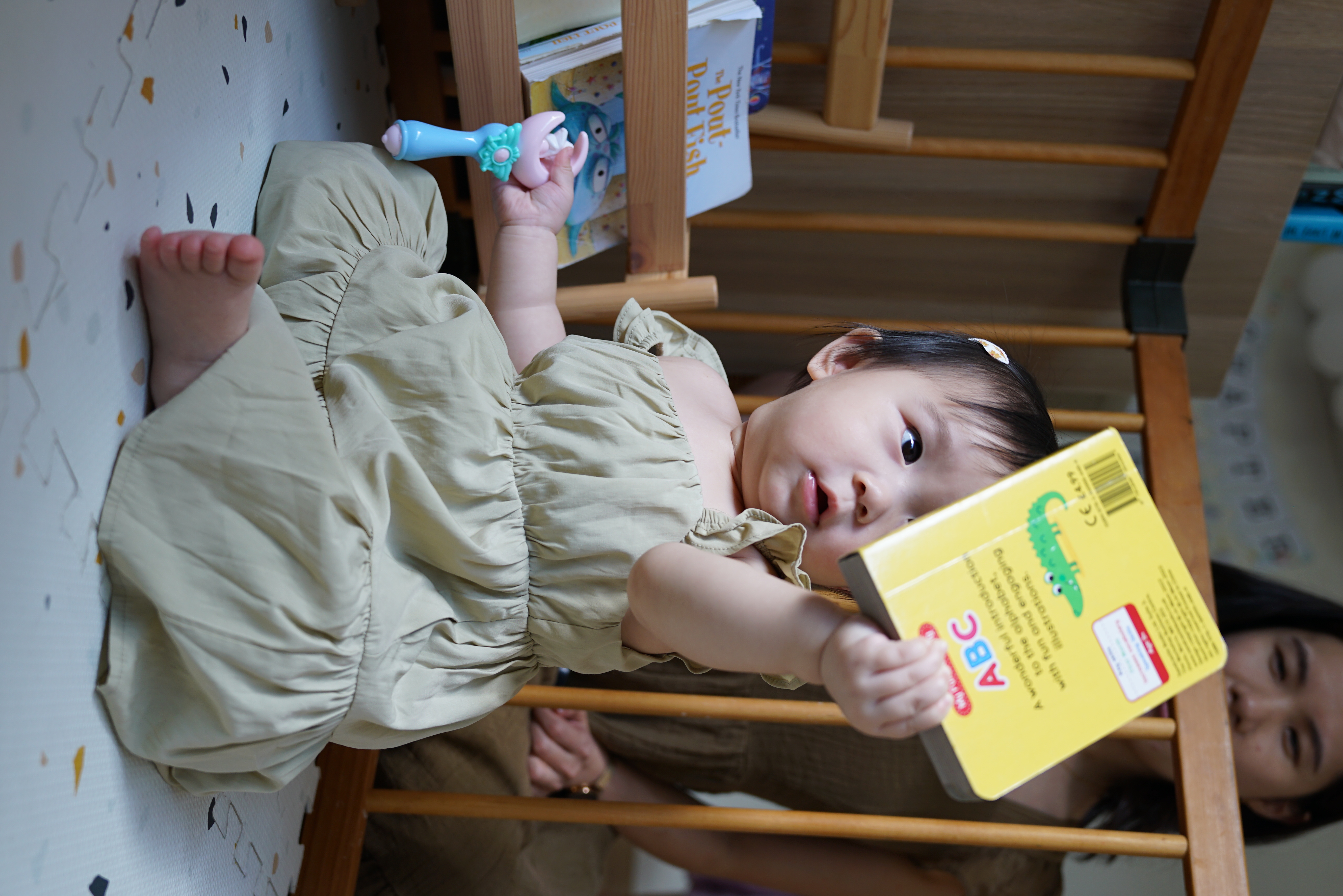 Reconfiguring the space for my toddler - Baby lying on bed, holding a book, wearing a beige dress, looking at the camera