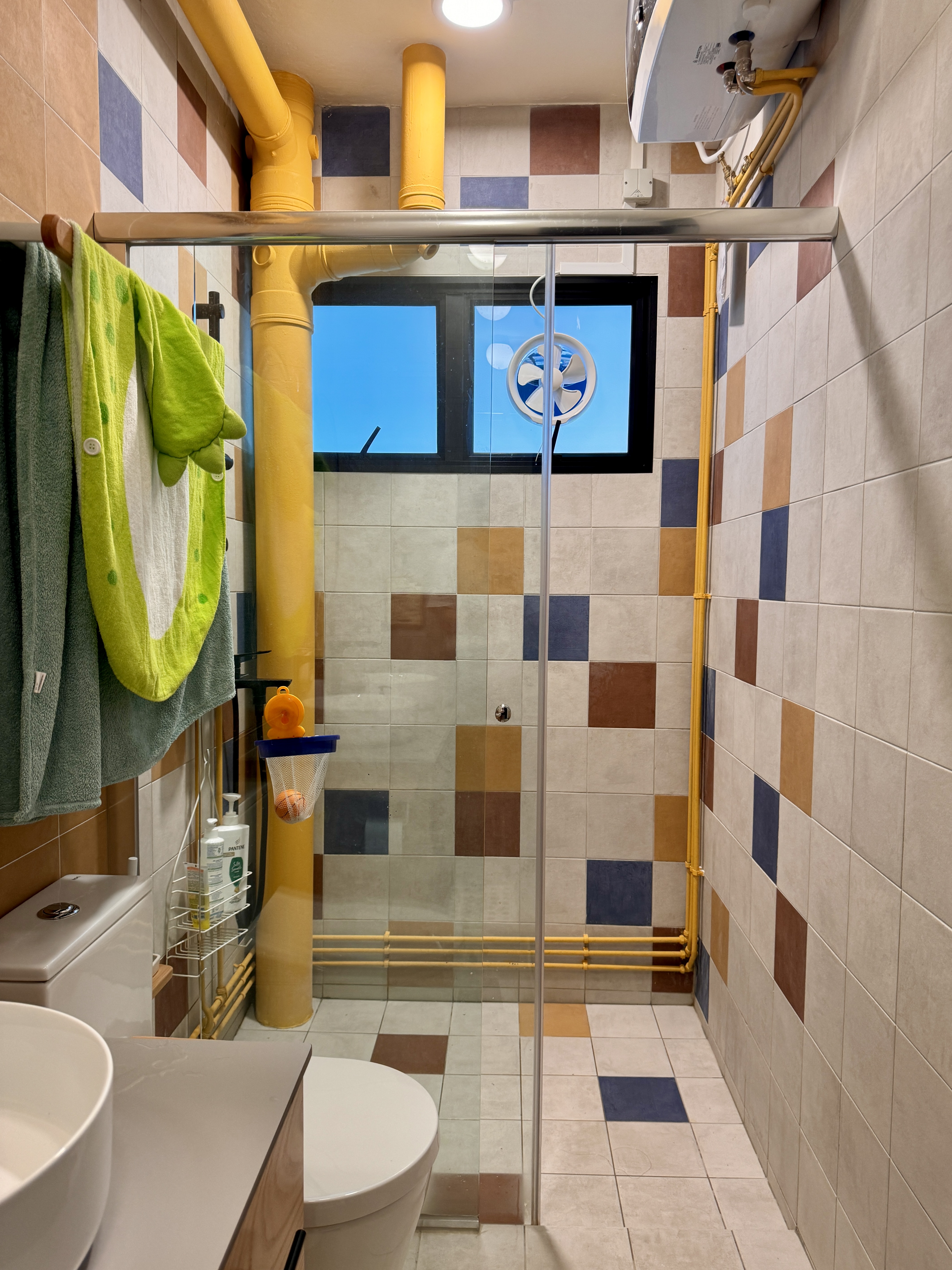 The Cheerful Swimming Pool (Bathroom 3/3) - Colorful tiled walls, yellow pipes, green towel, glass shower enclosure, blue and