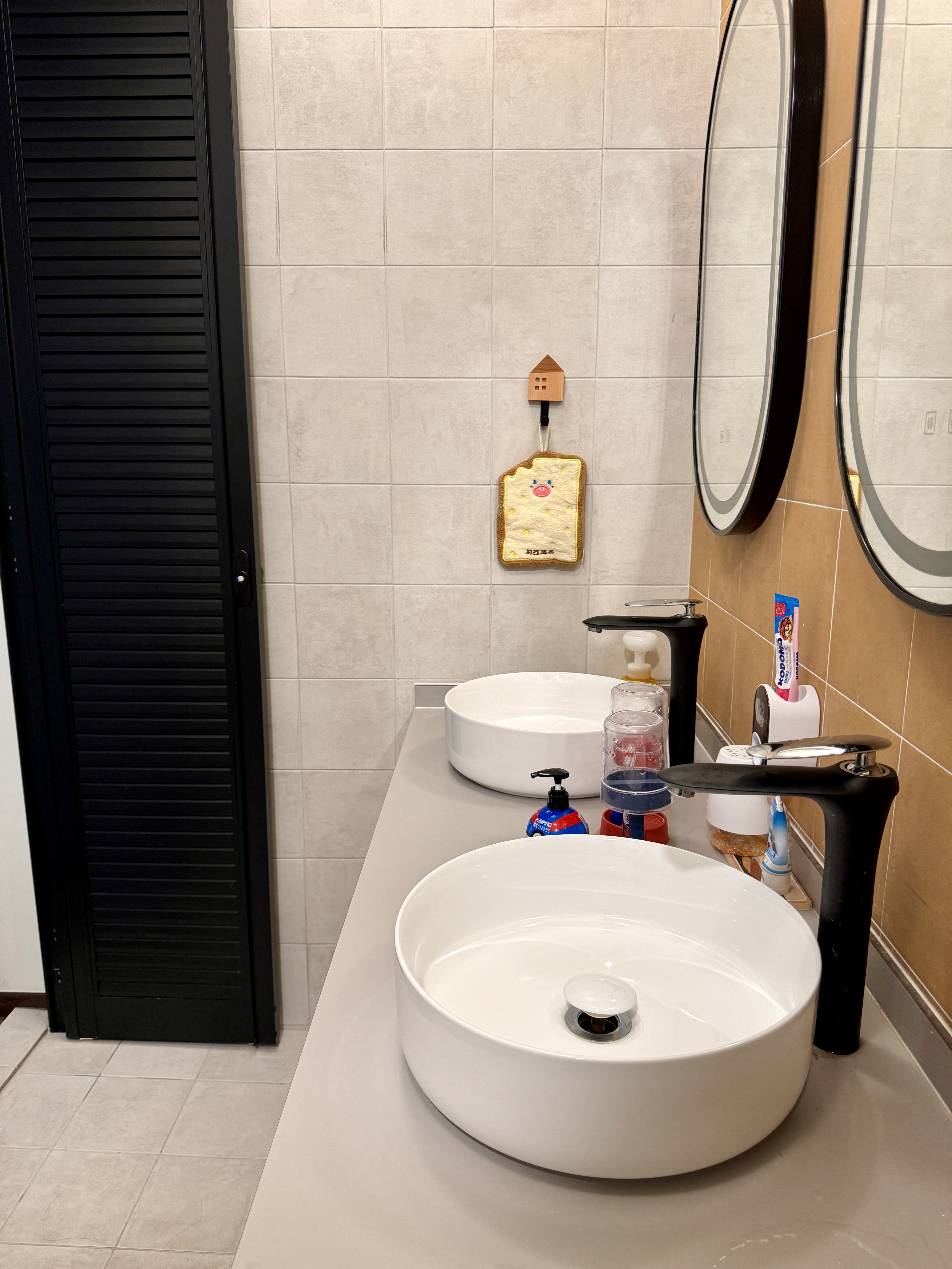 The Cheerful Swimming Pool (Bathroom 3/3) - White oval sinks, beige tiled walls, black bathroom cabinet, round mirror, black
