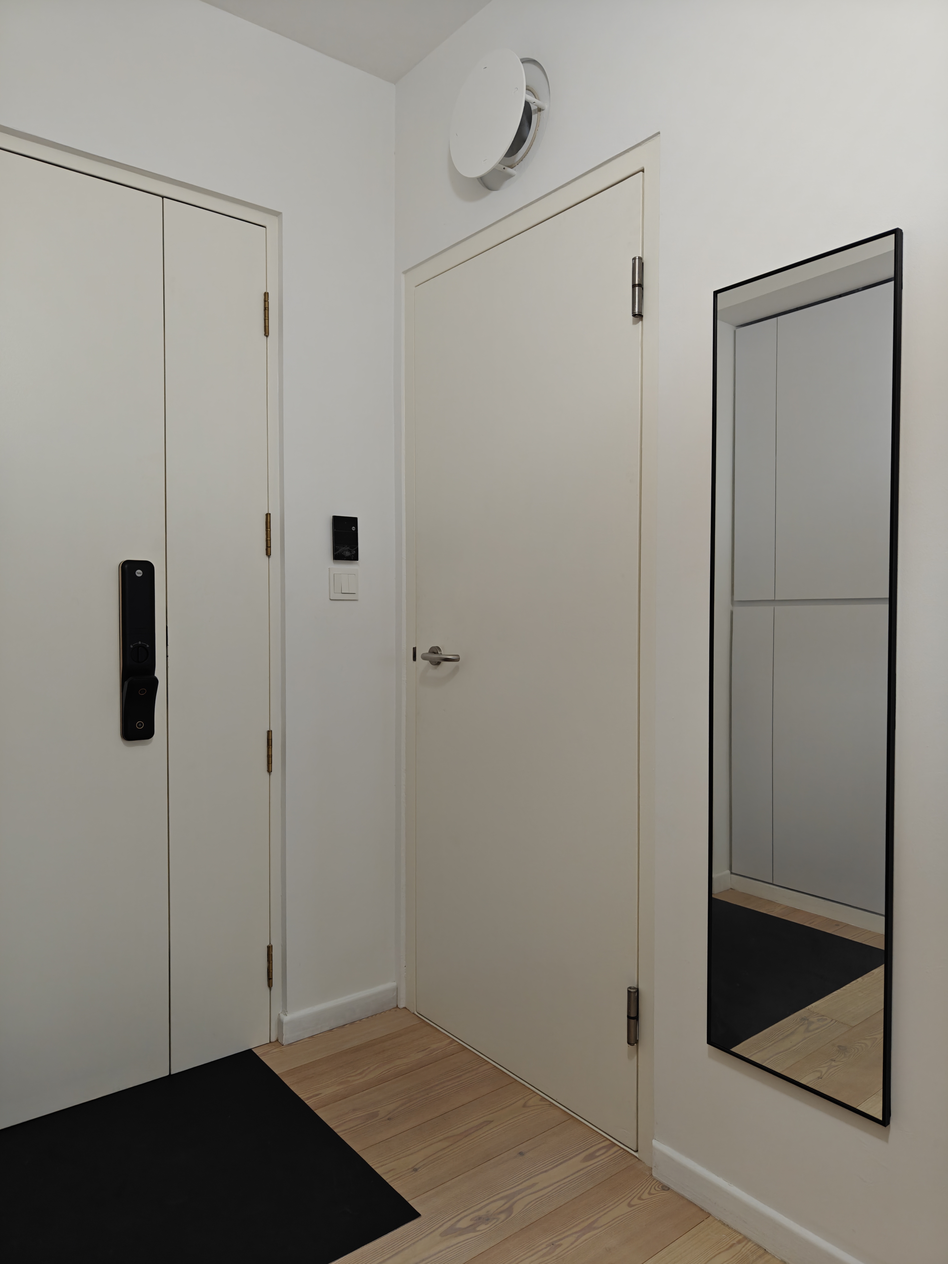 The Entry To Our World - White door with black handle, black electronic lock, beige wall, wooden floor, modern mirror