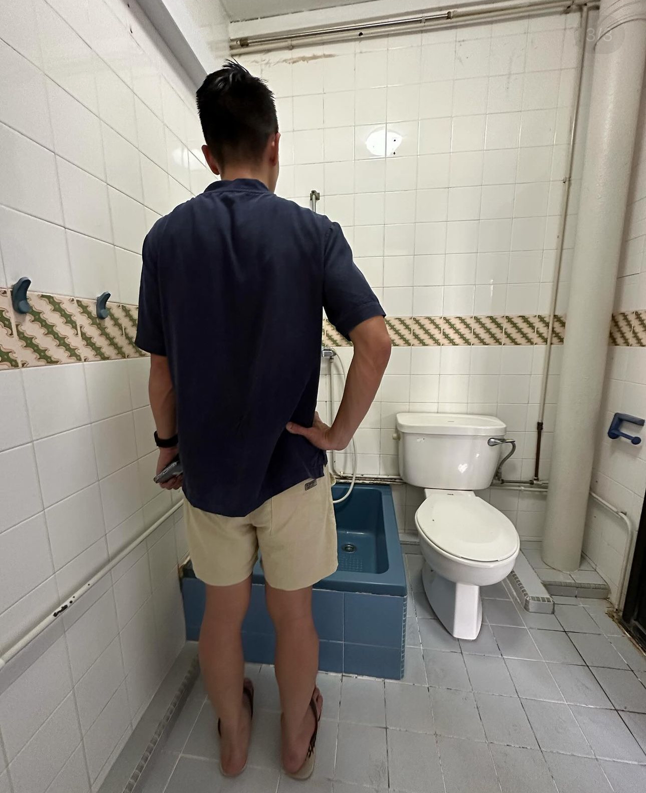 Join us in building our 3 bathrooms! - Bathroom, man standing, dark blue shirt, beige shorts, blue bathtub, white toilet