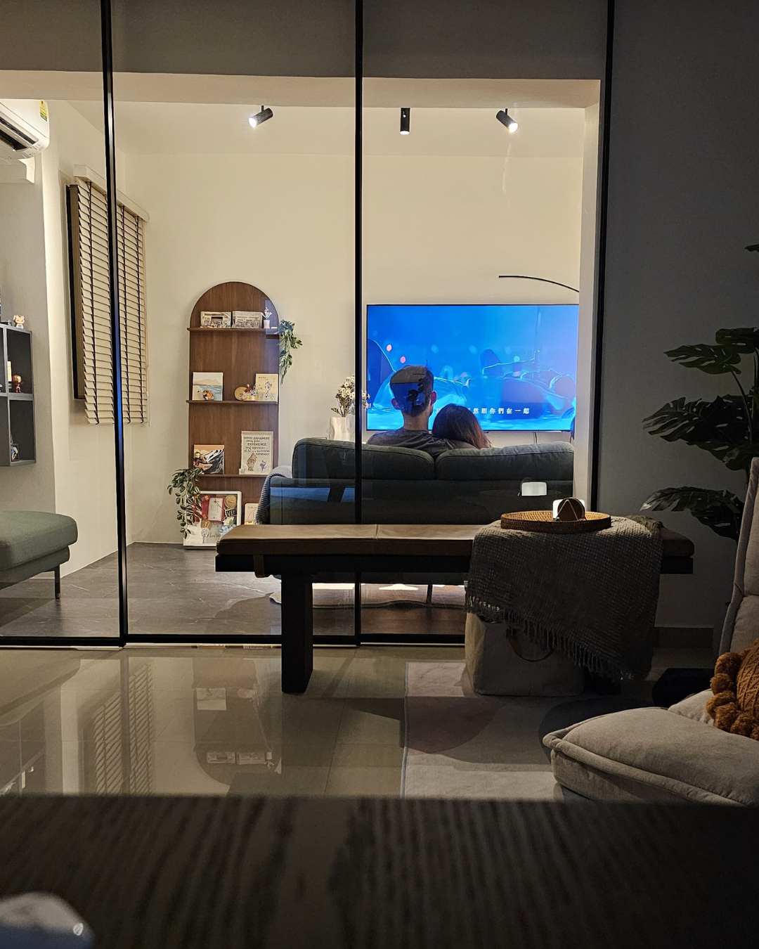 Hosting at Home: From 2 to 22  - Modern living room, cozy seating, glass wall, TV on, two people watching, indoor plants