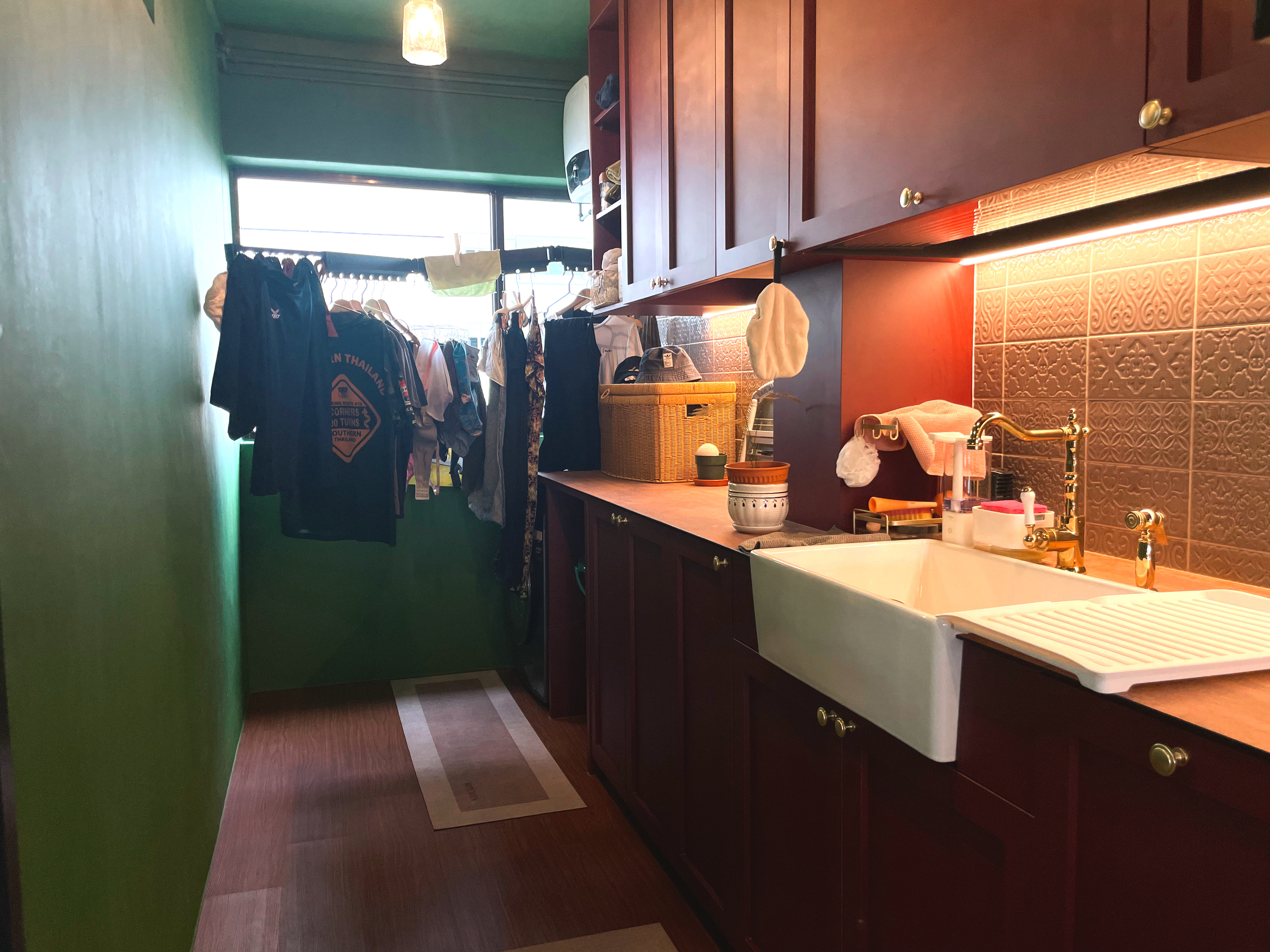 A No-Nonsense Laundry Corner - Green walls, wooden floor, hanging clothes, white sink, gold faucet, brown patterned tiles