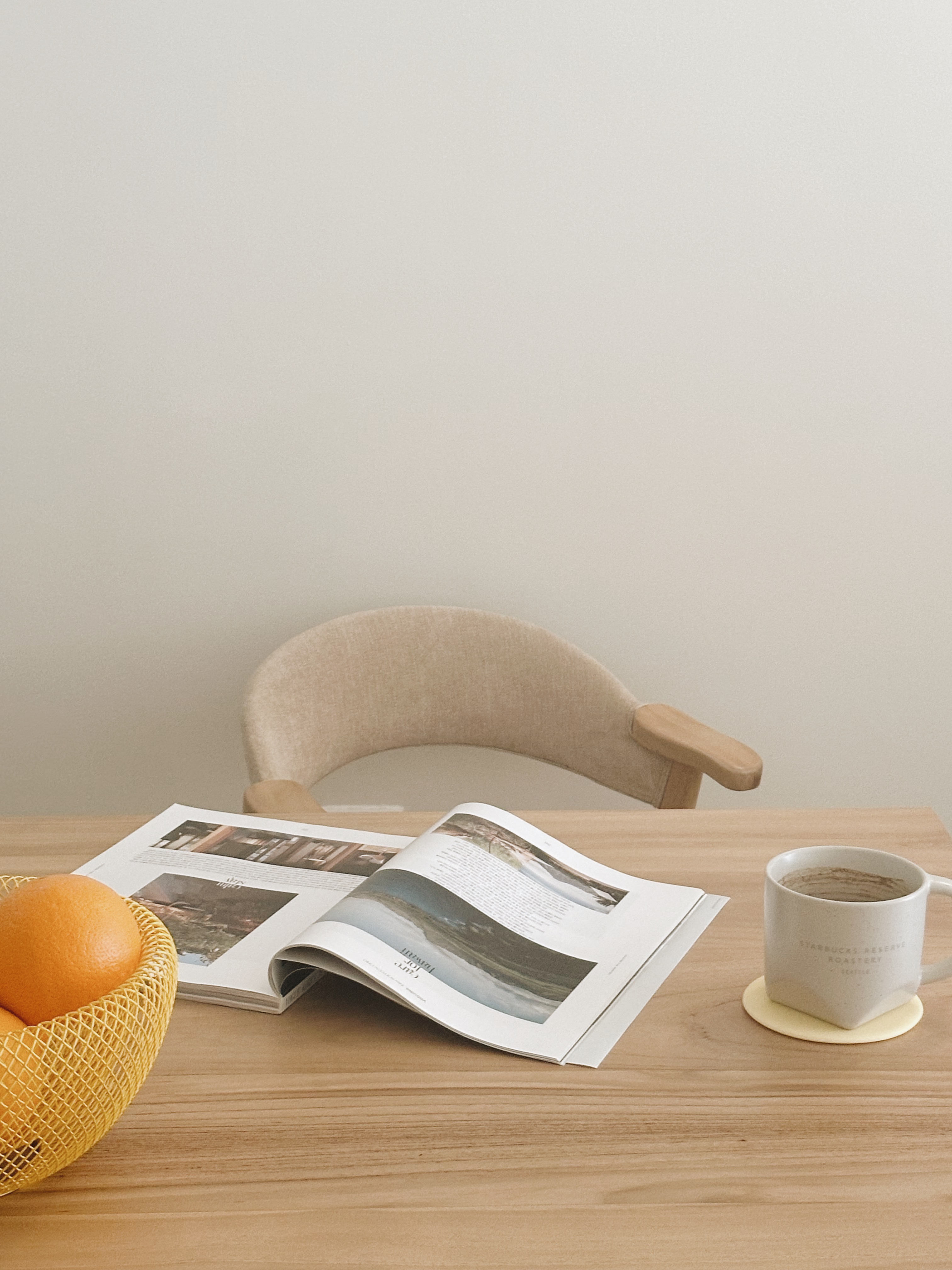 Inside a Warm Minimalist 3-Bedder Condo - Wooden table, open magazine, orange in basket, light beige chair, cozy room