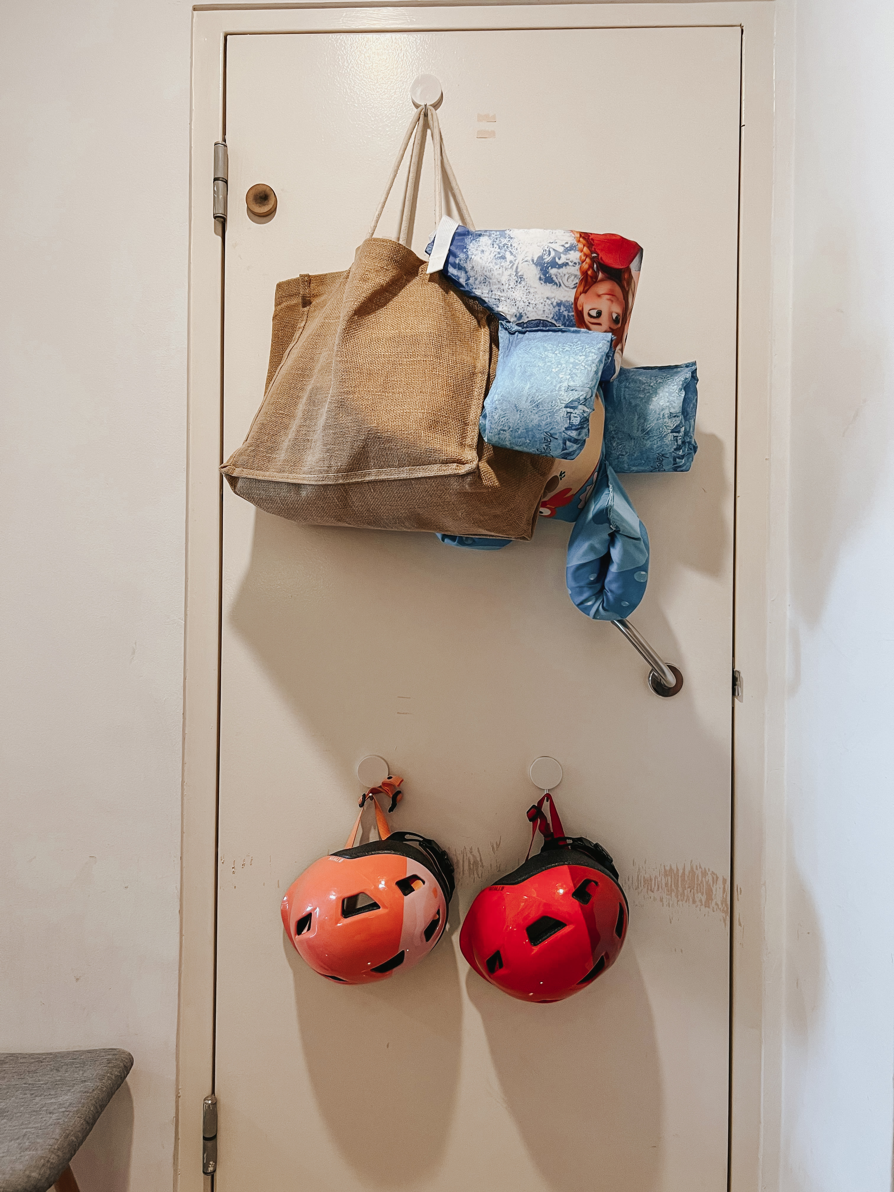 Our Entryway: A Practical Setup for Busy Families - Bags hanging on hooks, colorful helmets, beige door, small stool, orange