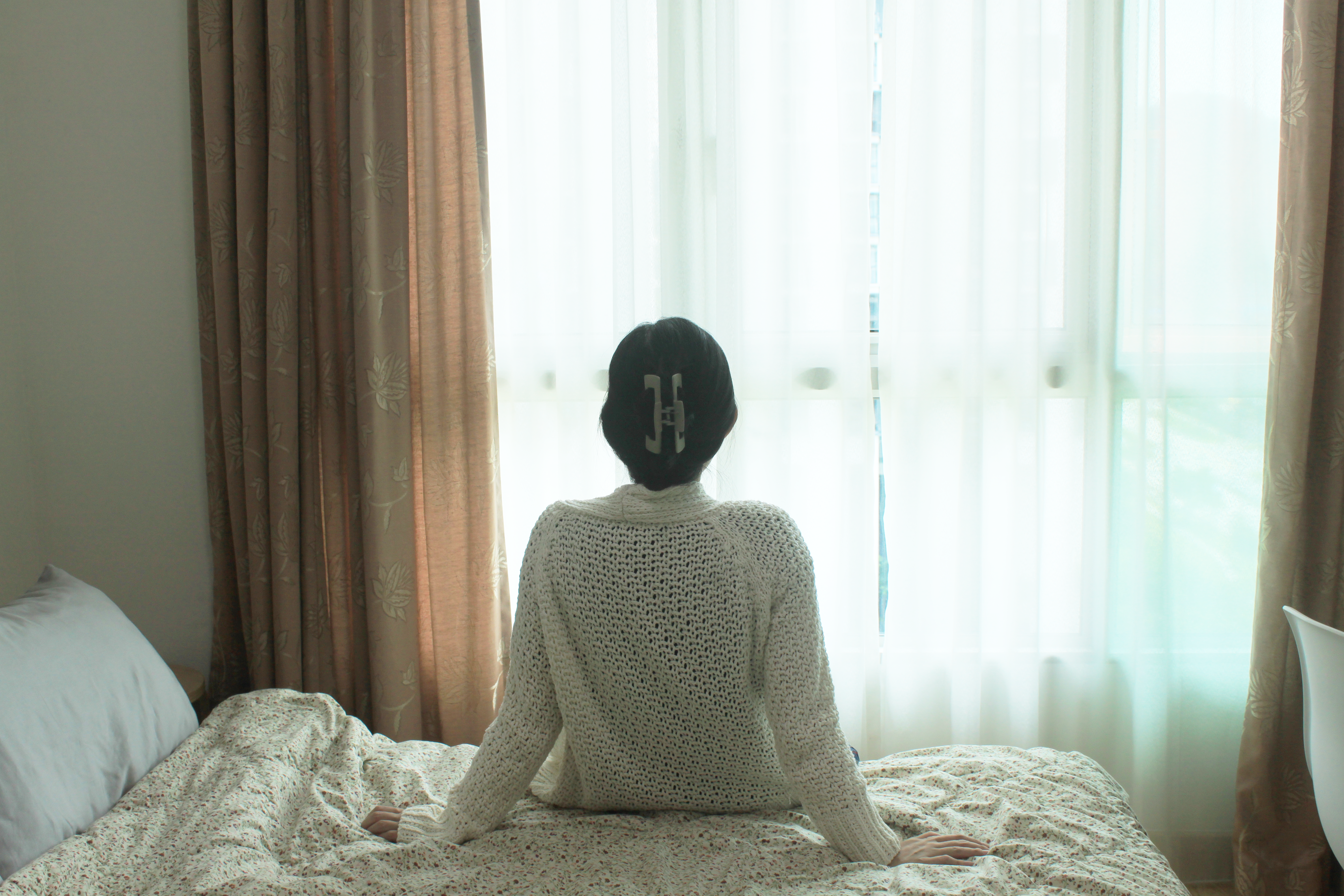 My Home Away From Home - Light streaming through sheer curtains, person sitting on bed, white floral bedspread, beige
